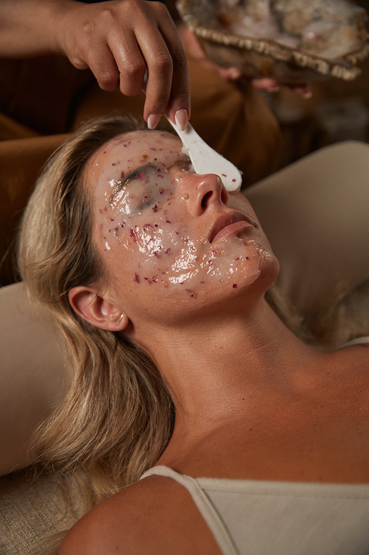 A guest mid-facial, product on the skin, the esthetician’s hand at her temple.