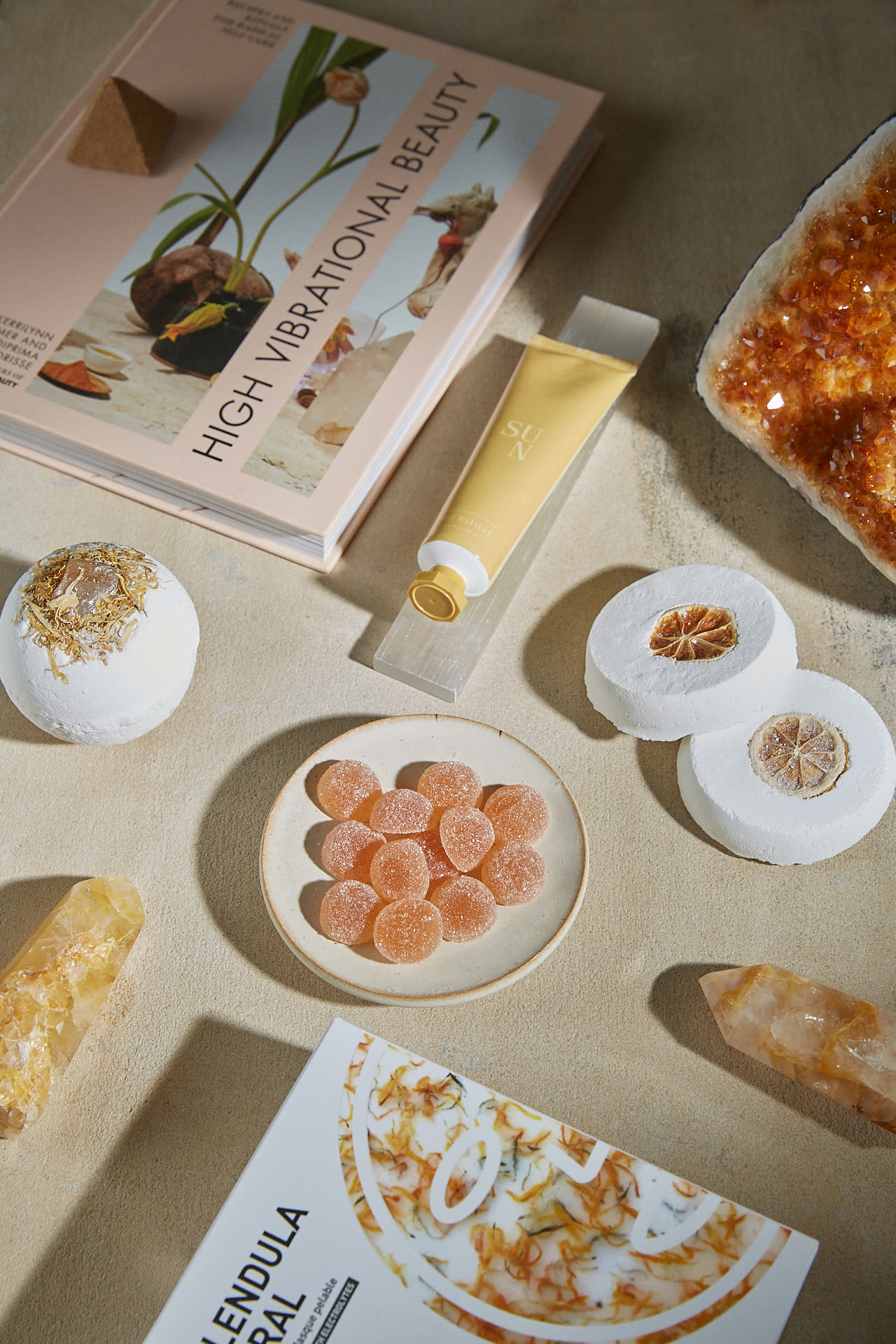 A flat-lay from the Edit: a book on high vibrational beauty, a gold-capped tube, bath fizzes, apricot pastilles and raw citrine and quartz points on linen.
