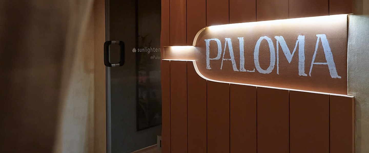 The threshold to the PALOMA sauna, the wordmark lit from within the clay-coloured wall.